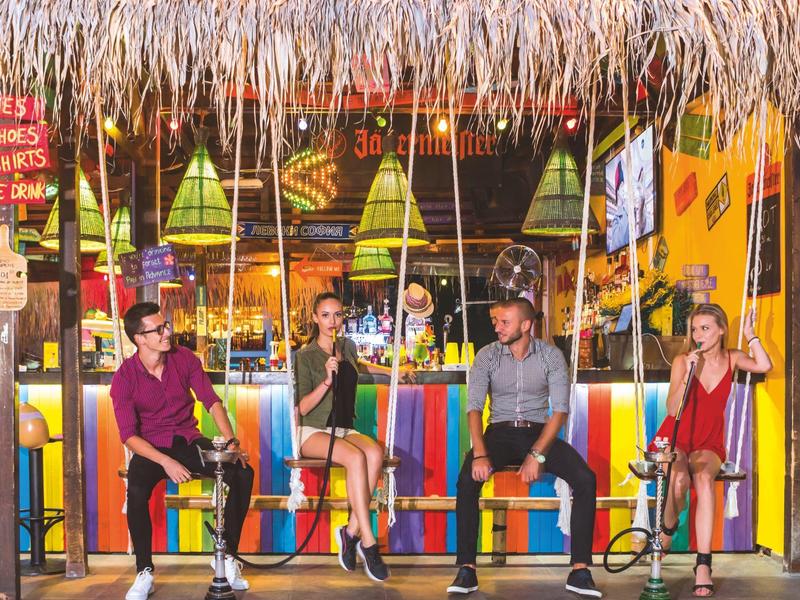 Vier Personen sitzen an einem farbenfrohen Barbereich mit Strohdach und rauchen Shisha.