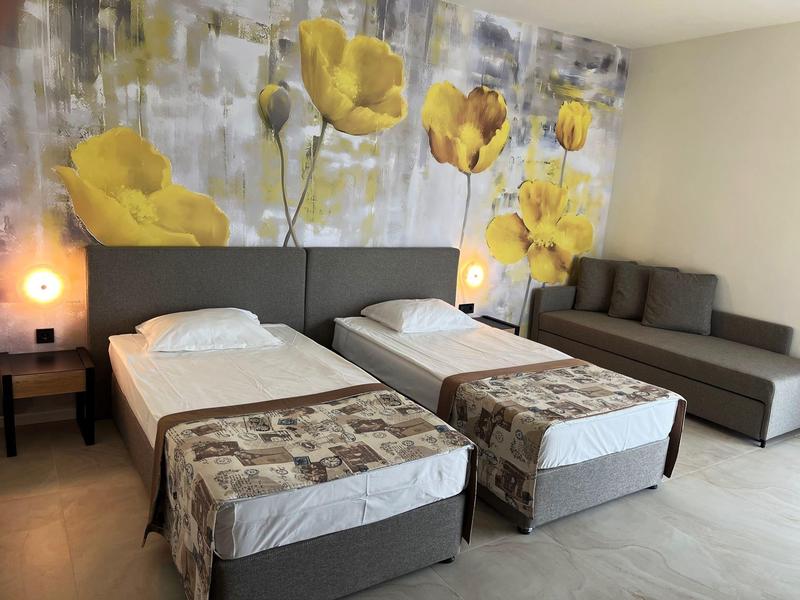 Zwei Einzelbetten mit grauen Kopfteilen vor einer Wand mit gelben Blumendekor, daneben ein graues Sofa.