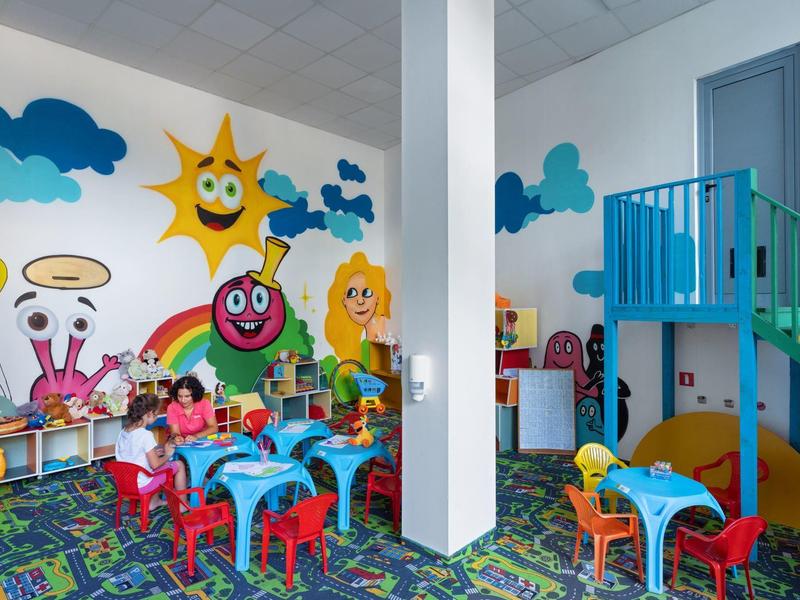 Bunter Kinderspielraum mit kleinen Tischen, Stühlen und Wandmalereien von Sonne und Figuren.