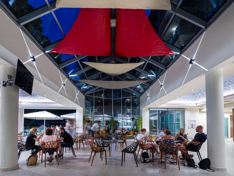 Modernes Hotelrestaurant mit geöffnetem Dach, roten Segeln und Gästen an Tischen