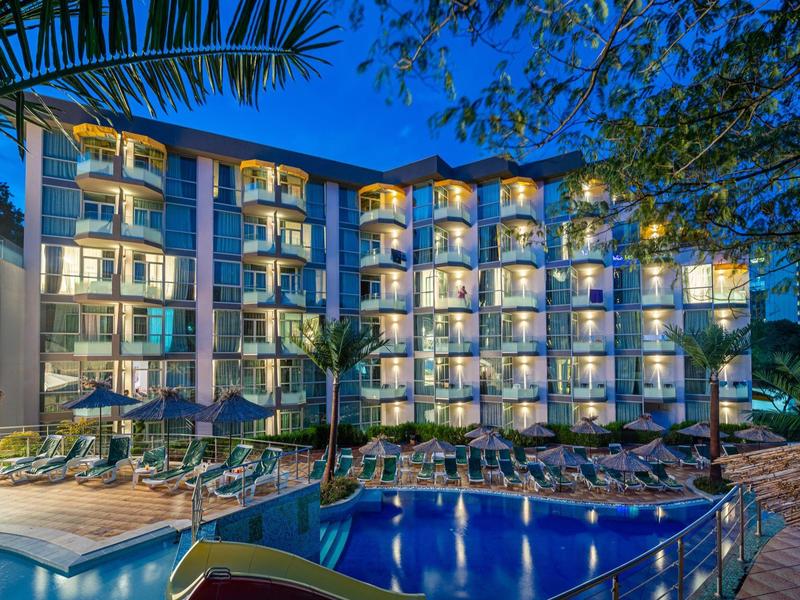 Modernes Hotel mit beleuchtetem Pool, Liegestühlen und Balkonen in tropischer Umgebung bei Abenddämmerung.