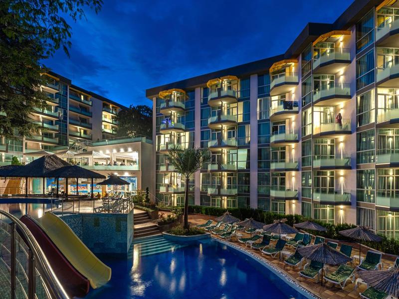 Beleuchtetes Hotel mit Balkonen und Pool bei Nacht, umgeben von Sonnenliegen und Rutschen.