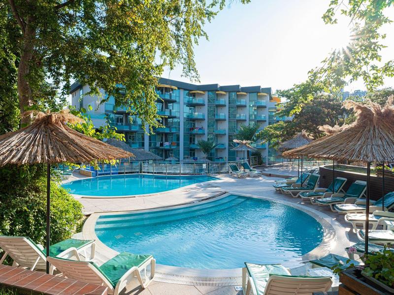 Großer Hotelpool mit Sonnenliegen und Strohdächern, umgeben von Bäumen und Hotelgebäude.