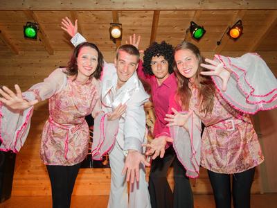 Vier Personen in farbenfrohen Kostümen posieren fröhlich in einem Holzraum mit Bühnenlicht.