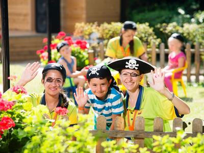 Drei lächelnde Kinder mit Piratenhut und Sonnenbrillen hinter einem Gartenzaun im Freien.