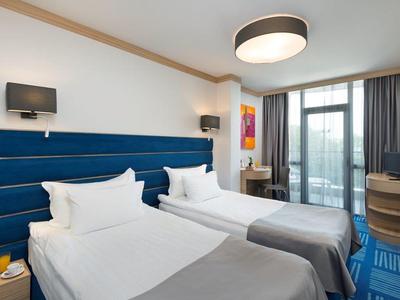 Moderne hotelkamer met twee eenpersoonsbedden, blauw hoofdeinde, bureau en groot raam met gordijnen.