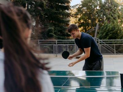 Zwei Personen spielen Tischtennis im Freien neben Bäumen und einem Geländer.