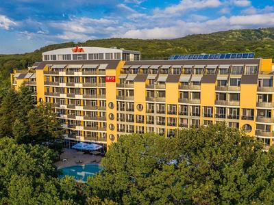 Groot hotelgebouw met gele gevel, omringd door groene bomen en blauwe lucht.