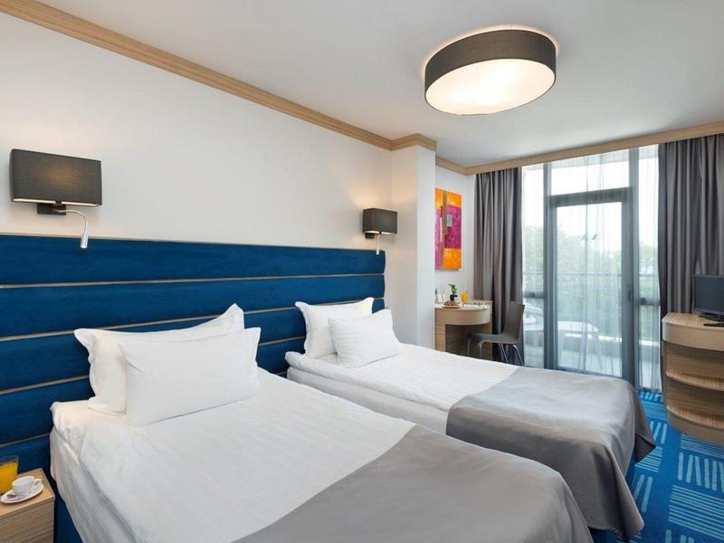 Moderne hotelkamer met twee eenpersoonsbedden, blauw hoofdeinde, bureau en groot raam met gordijnen.