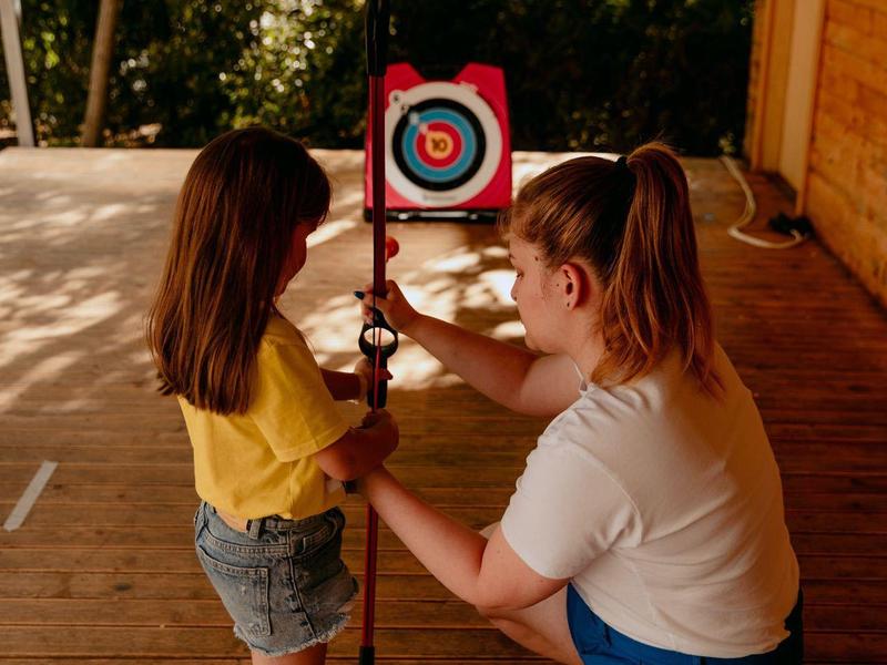 Een vrouw leert een meisje boogschieten buiten, beiden geconcentreerd op het doel.