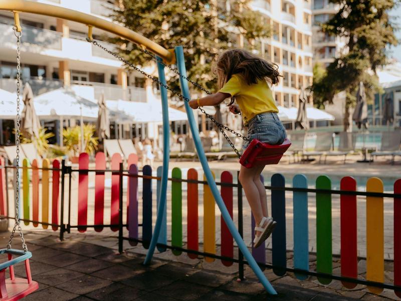Kind schommelt op een kleurrijke speeltuin naast een hotelgebouw.