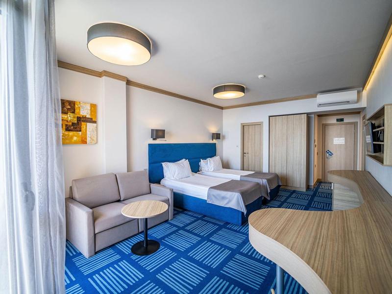 Modern hotelkamer met twee bedden, bank, bureau en lichtblauwe vloerbedekking.