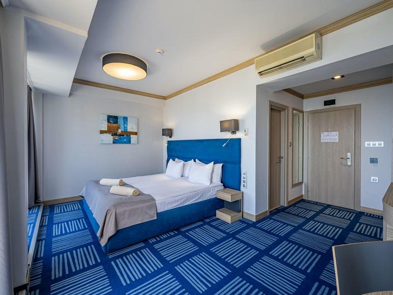 Modern hotelkamer met blauw tapijt, tweepersoonsbed en bureau in daglicht.
