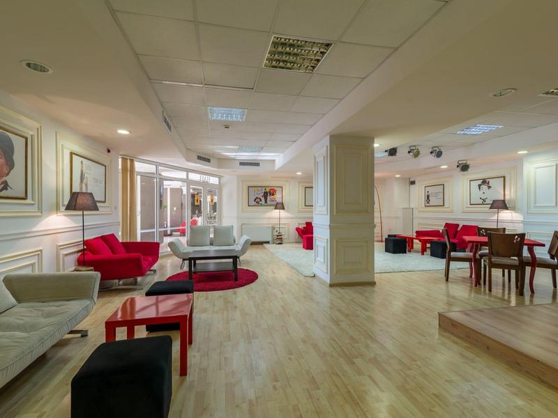 Área de salón de hotel moderna y luminosa con suelo de madera, muebles rojos y blancos, y arte en las paredes.