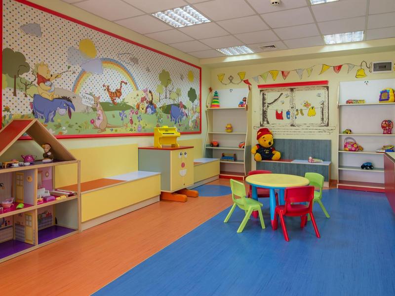 Sala de juegos infantil con mesa redonda, sillitas de colores y estanterías con juguetes y muñecos.