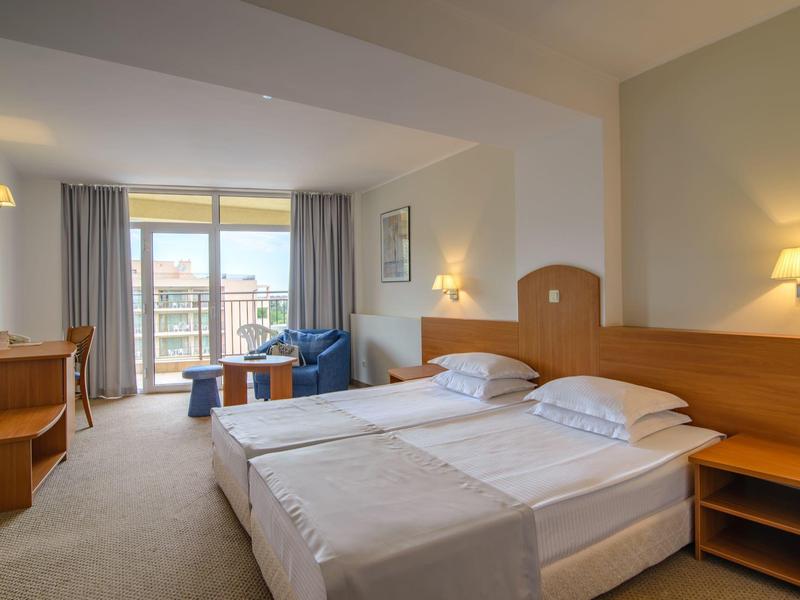 Habitación de hotel luminosa con dos camas, balcón y zona de estar con sillones azules.