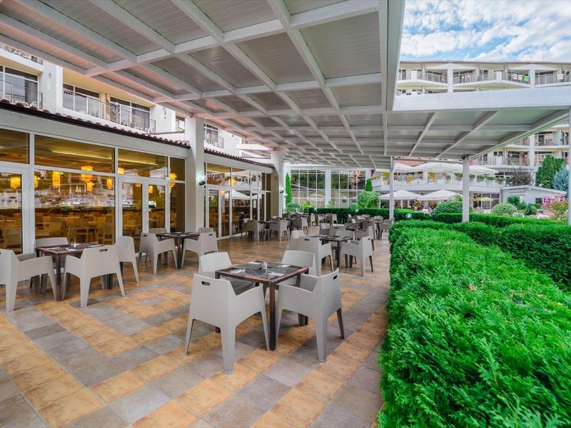 Zona exterior moderna de un hotel con mesas, sillas y setos verdes bajo techo.