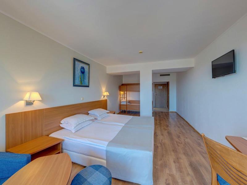 Habitación de hotel luminosa con cama doble, muebles de madera, televisor y área de estar con silla y mesa.