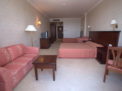 Hotelzimmer mit zwei Betten, rotem Sofa, Holztisch und Stuhl, beigem Teppich und Wandschmuck.
