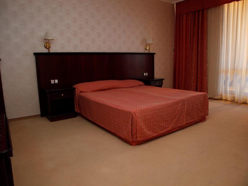 Schlichtes Hotelzimmer mit Doppelbett, braunem Kopfteil, Nachttischen und Fenster mit Vorhängen.