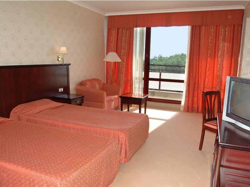 Zweibettzimmer mit rot-orange Bettwäsche, Vorhängen, Sessel, Holzmöbeln und Fernseher am Fenster.