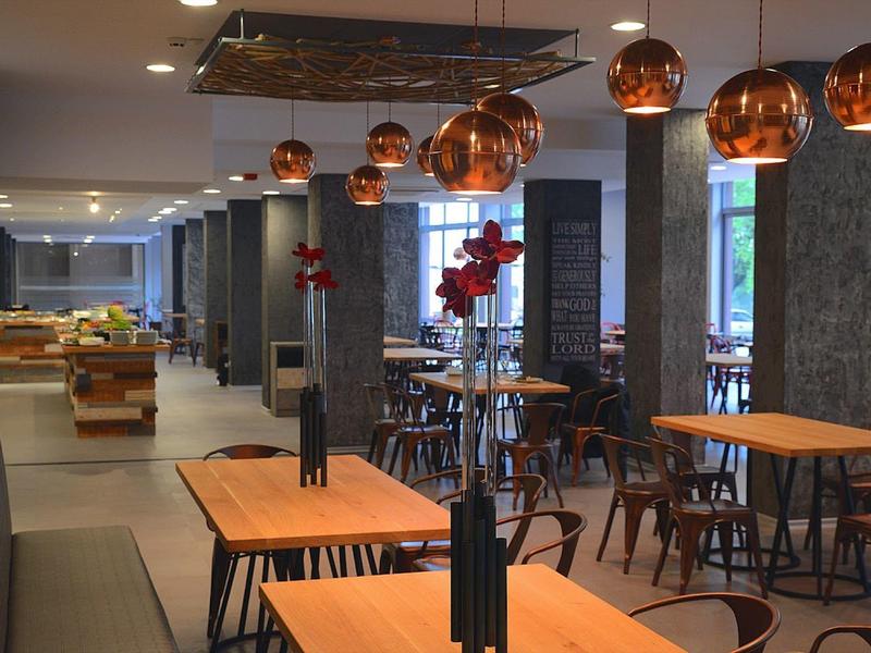 Modernes Restaurant mit Holztischen, kupfernen Hängeleuchten und großen Fenstern.