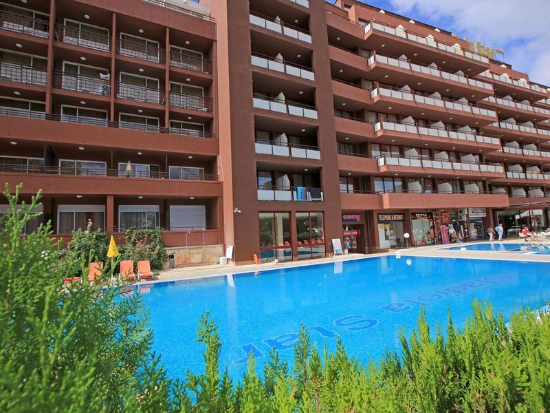 Großes Hotelgebäude mit mehreren Balkonen und klarem blauen Pool im Vordergrund, umgeben von Grün.