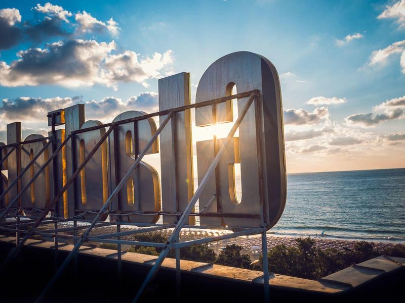 Große weiße Buchstaben auf einer Plattform am Meer, Sonnenlicht durchscheint, blauer Himmel mit Wolken.