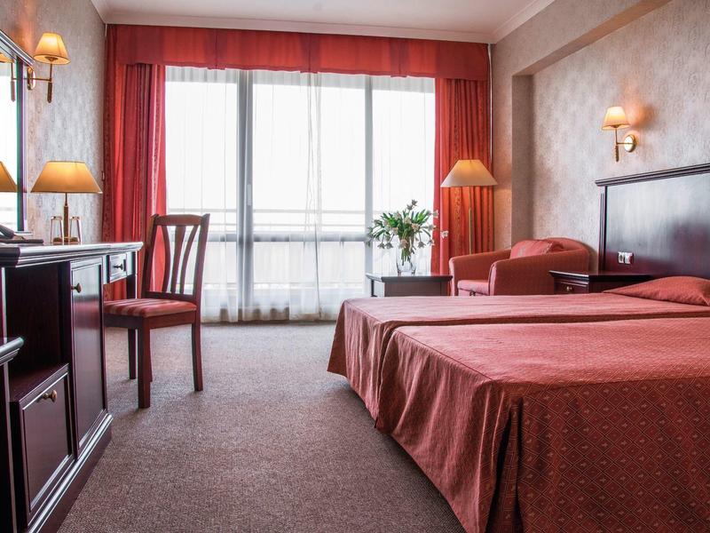 Zimmer mit Doppelbett, roten Vorhängen, Sessel, Schreibtisch und Teppichboden in warmen Farben.