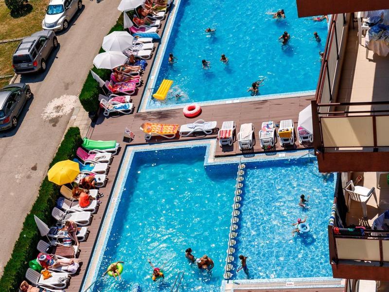 Zwei rechteckige Schwimmbecken mit vielen Menschen, Sonnenschirmen und Liegestühlen neben einem Gebäude.