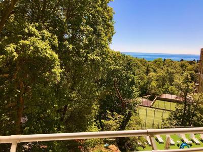 Blick von Balkon auf grüne Bäume und Tennisplatz, im Hintergrund das blaue Meer und klarer Himmel.