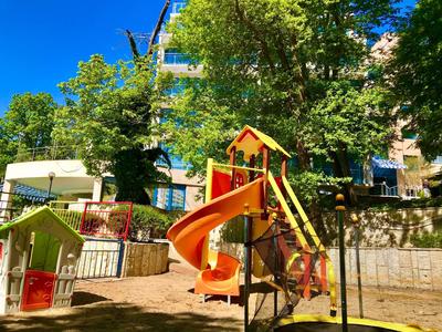 Kinderspielplatz mit orangefarbener Rutsche und Klettergerüst, umgeben von Bäumen und Gebäuden.
