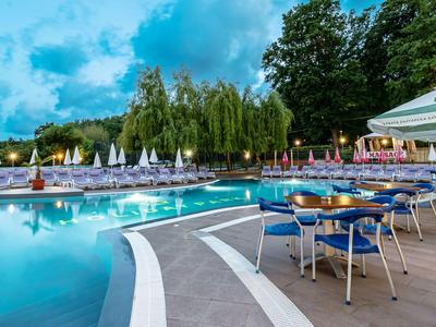 Abendansicht eines Pools mit leeren Stühlen und Tischen am Hotel, umgeben von Bäumen und Sonnenschirmen.