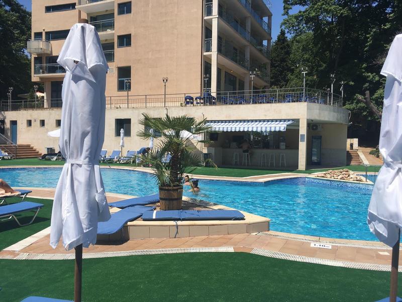 Außenpool mit Liegestühlen, weißen Sonnenschirmen, Palmen und einem mehrstöckigen Gebäude im Hintergrund.