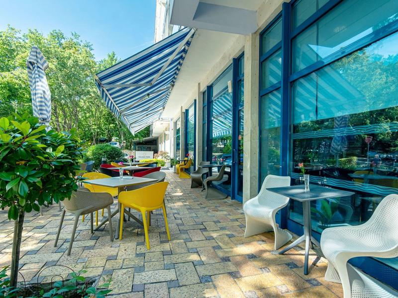 Moderne Terrasse mit bunten Stühlen, Glastisch und großem Glasfenster, umgeben von Grün.