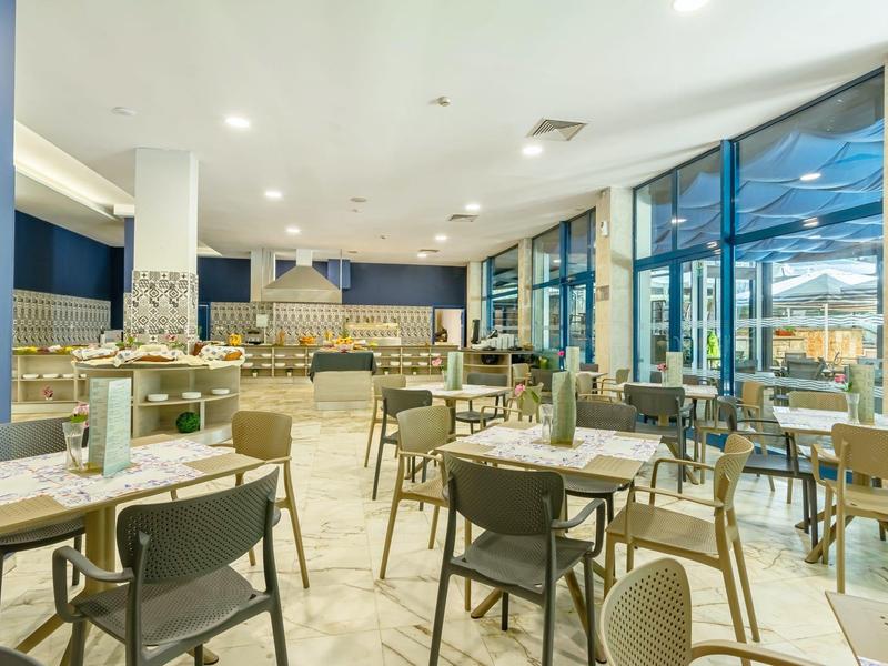 Modernes Restaurant mit grauen Stühlen, Holz- und Metalltischen, großen Fenstern und heller Beleuchtung.