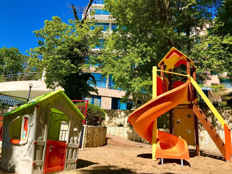 Bunter Kinderspielplatz mit Spielhaus und rutschiger Rutsche vor grünem Bewuchs und blauem Himmel.