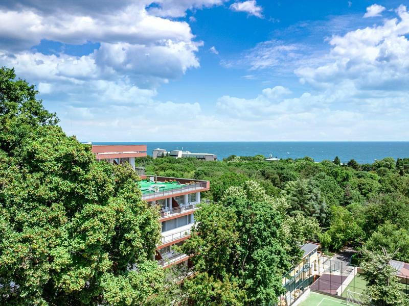 Grüne Bäume, modernes Gebäude und blauer Himmel mit Wolken über dem Meer im Hintergrund.