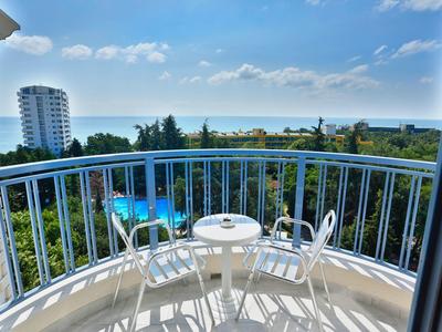 Balcón con mesa y sillas, vista al mar, árboles y edificios bajo cielo soleado.