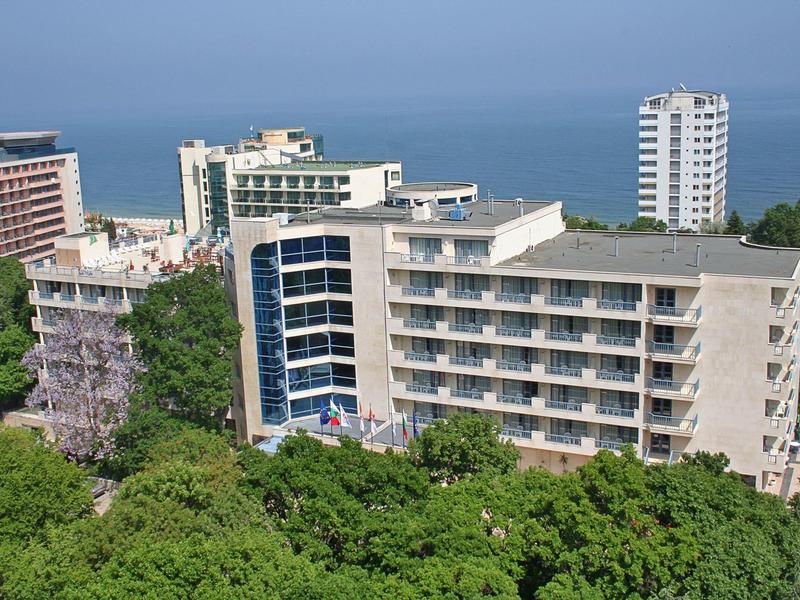 Edificio de hotel de varios pisos junto al mar, rodeado de abundante vegetación y otros edificios.
