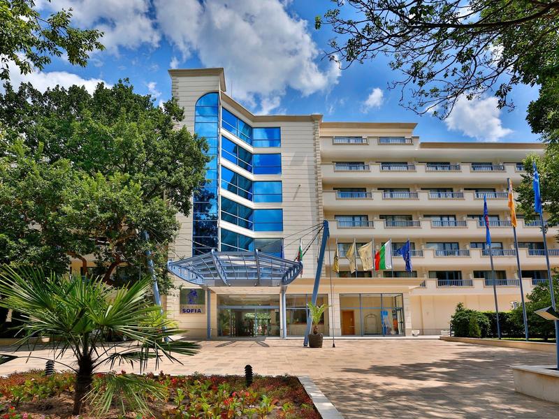 Complejo hotelero moderno con entrada de varios pisos, árboles y cielo azul de fondo.