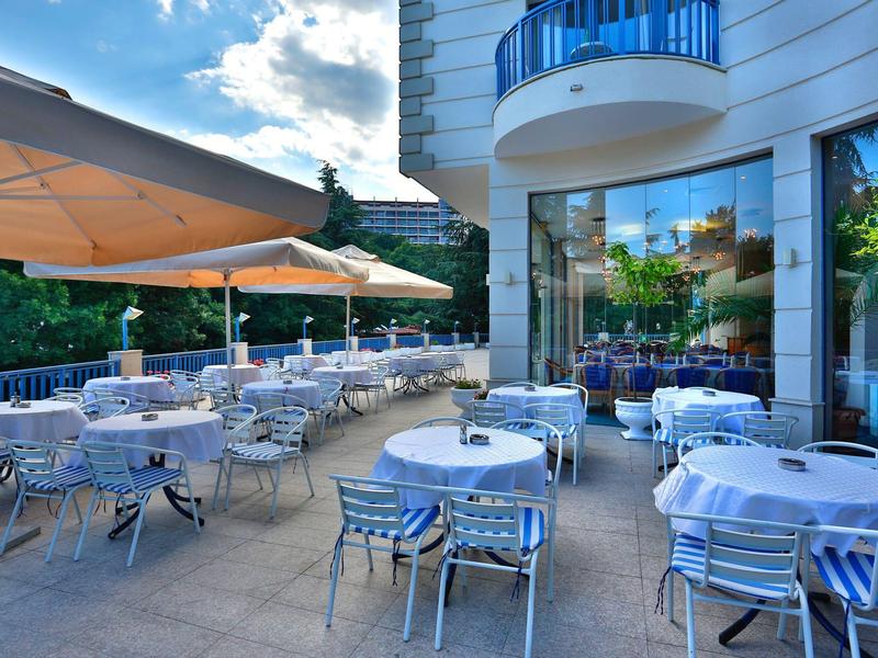 Restaurante al aire libre con mesas redondas, manteles blancos y sillas de metal junto a un hotel.