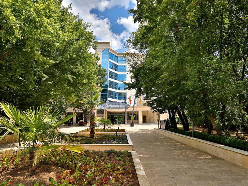 Camino conduce a edificio hotelero moderno, rodeado de árboles y áreas de jardín bajo cielo azul.