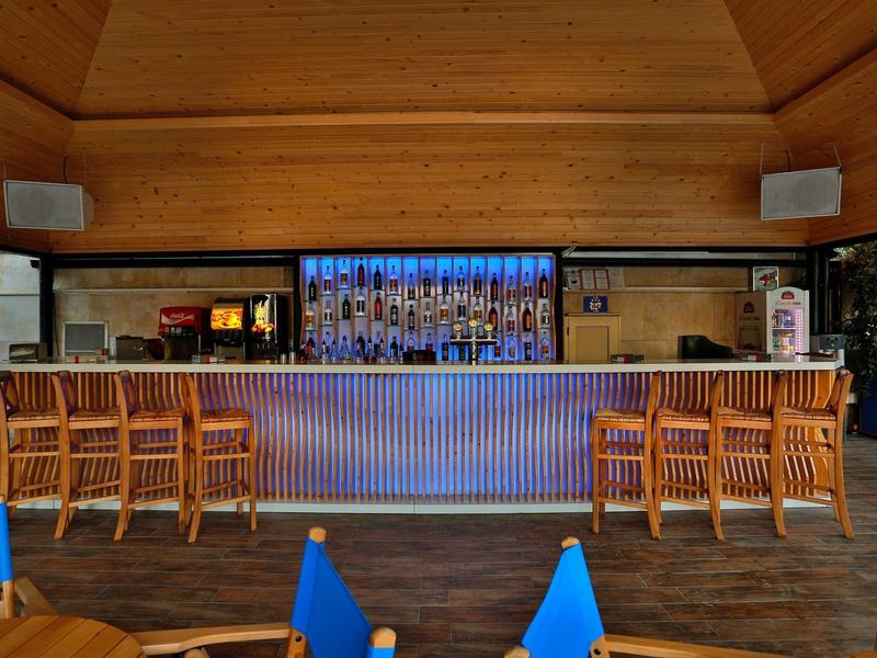 Área de bar moderna de hotel con mostrador de madera, taburetes y pantalla grande al fondo.