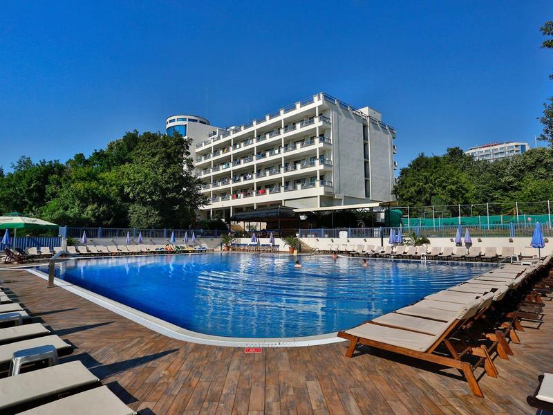 Gran hotel con piscina exterior, rodeado de tumbonas y árboles bajo un cielo despejado.