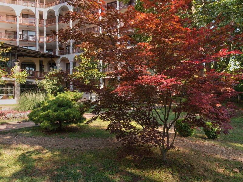 Giardino autunnale con alberi colorati davanti a un edificio alberghiero su più piani.