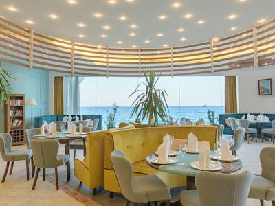 Helles Hotelrestaurant mit Panoramafenstern, gelbem Sofa und Blick auf das Meer.
