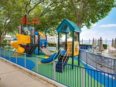 Ein bunter Kinderspielplatz mit Rutschen neben einem Bereich mit Sonnenschirmen und Sitzgelegenheiten am Wasser.