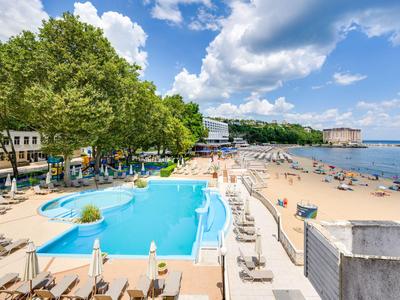 Hotel mit Pool und Strandbereich, umgeben von Liegestühlen und grünen Bäumen bei sonnigem Wetter.