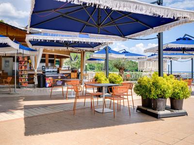 Außenbereich eines Cafés mit Tischen, Stühlen und großen blauen Sonnenschirmen in sonniger Umgebung.
